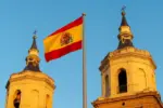 Aliança Evangélica Espanhola lança rede de apoio habitacional para famílias em situação de vulnerabilidade 6 Bandeira da Espanha entre as torres de uma igreja Foto Folha Gospel Canva IA